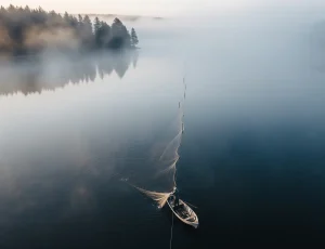 Ảnh chụp từ trên cao (drone view) cảnh một người đánh cá trên chiếc thuyền nhỏ đang giăng lưới rê trên mặt hồ tĩnh lặng, bao phủ bởi lớp sương mù huyền ảo vào buổi sớm.