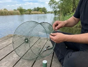 Cận cảnh đôi tay người đàn ông đang tỉ mỉ dùng kim và dây cước màu xanh để vá lại lỗ thủng lớn trên một chiếc lồng lưới tròn đựng cá. Bên cạnh trên sàn gỗ là kìm và cuộn dây cước, bối cảnh bờ sông