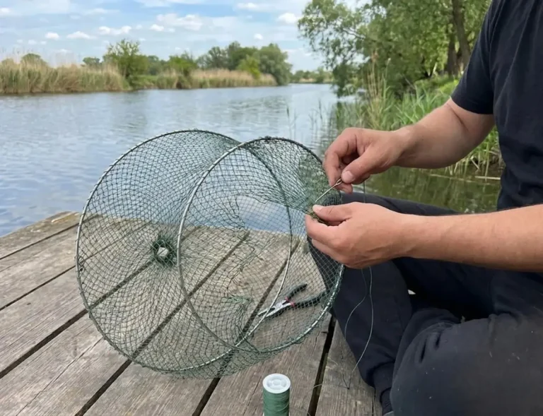 Cận cảnh đôi tay người đàn ông đang tỉ mỉ dùng kim và dây cước màu xanh để vá lại lỗ thủng lớn trên một chiếc lồng lưới tròn đựng cá. Bên cạnh trên sàn gỗ là kìm và cuộn dây cước, bối cảnh bờ sông